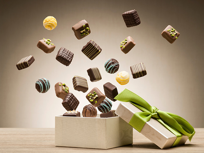 Open box with lid on the side wrapped in a green silk ribbon, exploding into the air from the box are more than a dozen small decorated chocolates.